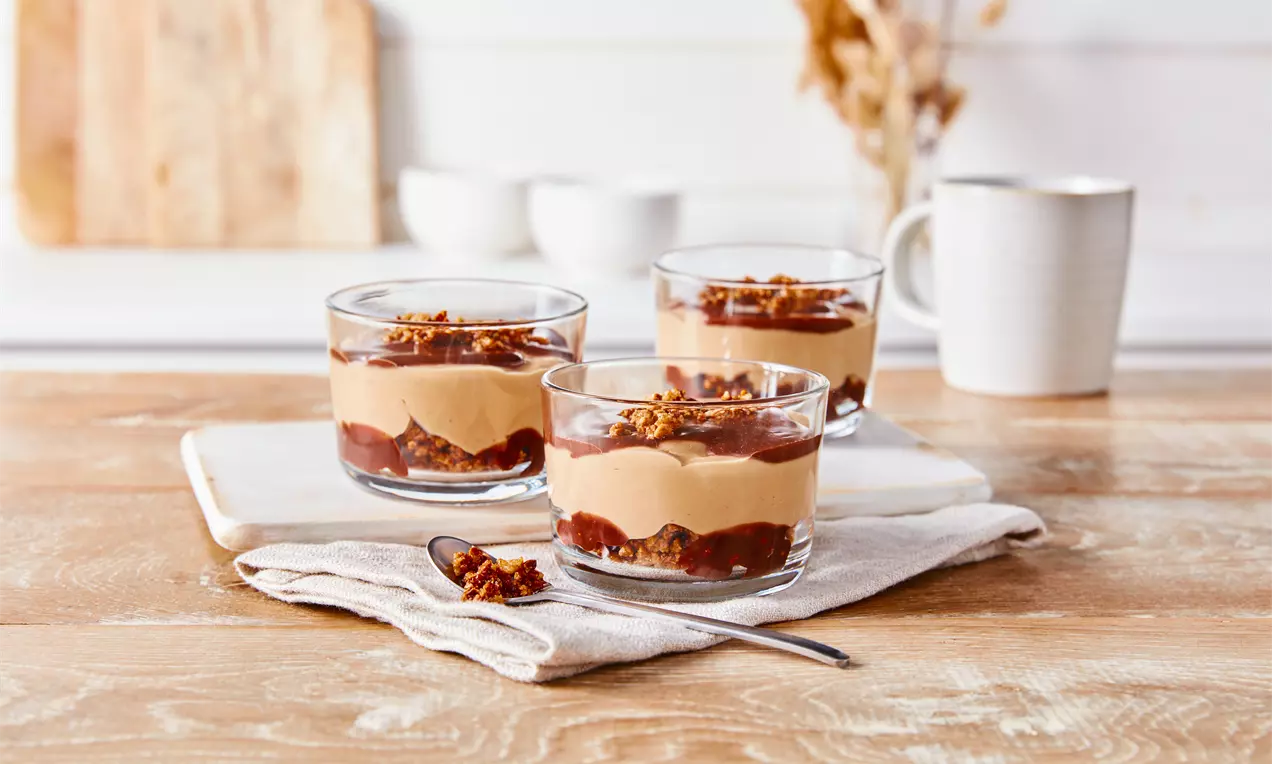 Budincă caramel stratificată în pahare transparente, cu straturi de cremă caramel aurie, fructe uscate și topping crocant pe o tavă albă